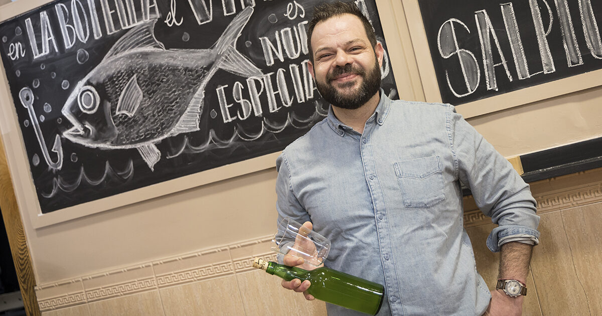 Pablo Fernández sonriendo con botella de sidre frente a pizarra con dibuxu de pexe nel chigre La Botella n’Avilés