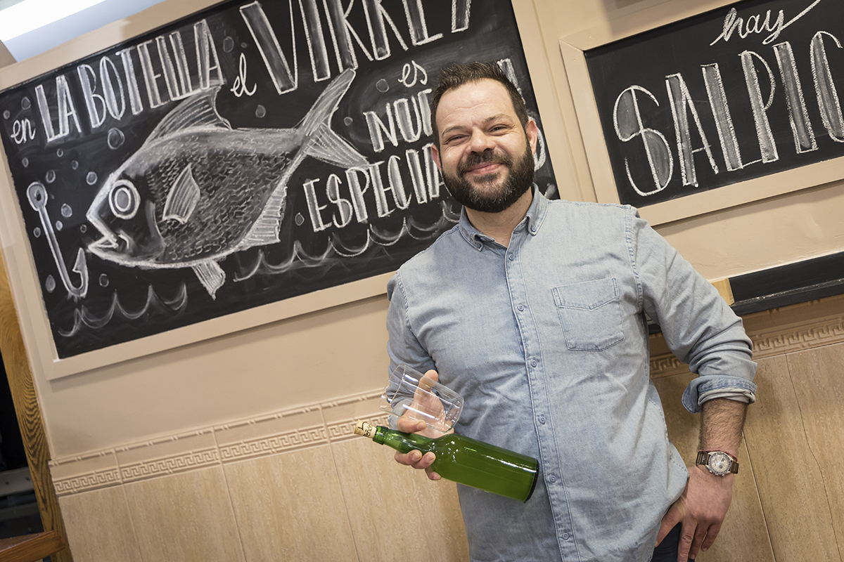 Pablo Fernández sonriendo con botella de sidre frente a pizarra con dibuxu de pexe nel chigre La Botella n’Avilés