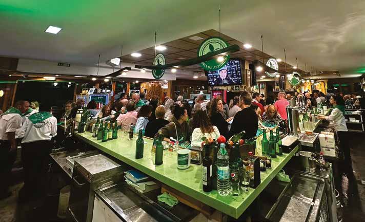 Personas alrededor de la barra de una sidrería, con botellas y vasos de sidra.